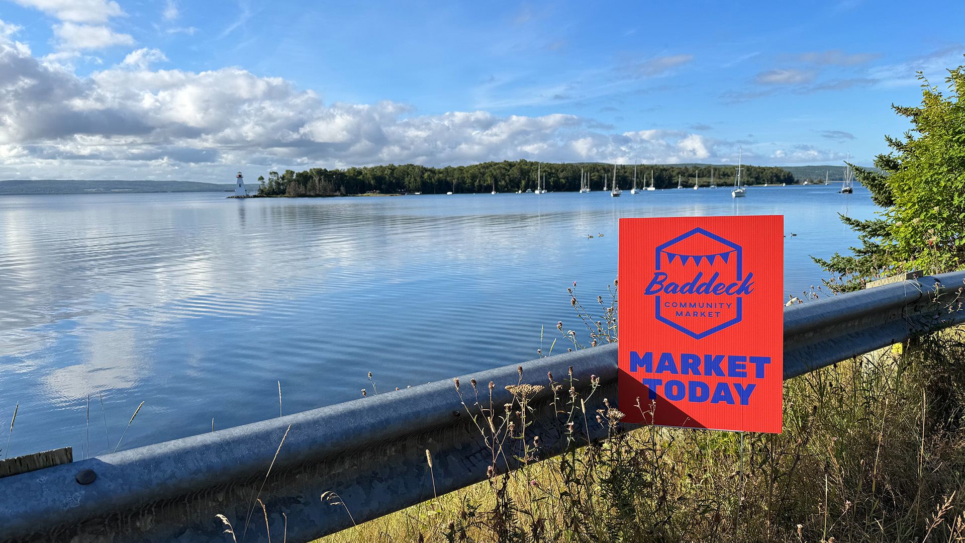 Baddeck Community Market sign by the waterfront, with sailboats and the Kidston Island Lighthouse in the distance.